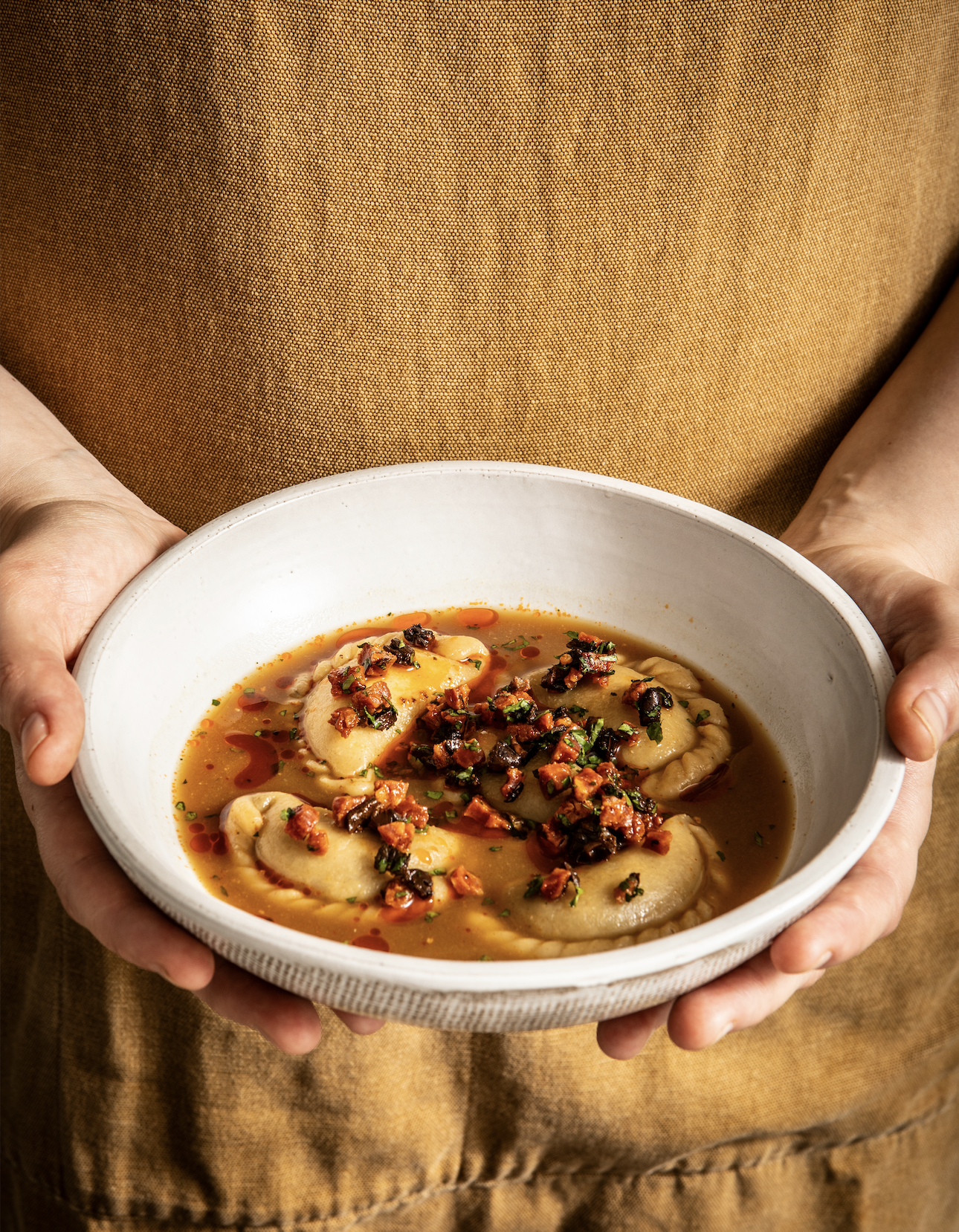 A person holding a white bowl of dumplings in broth, topped with chopped vegetables and herbs.