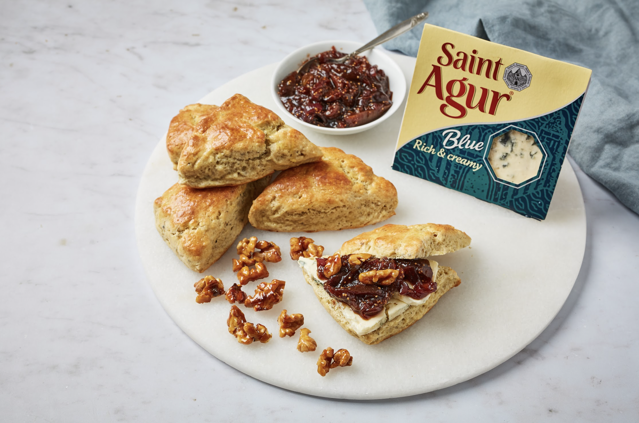 Scones with cheese and jam, a bowl of jam, and a box of Saint Agur Blue cheese on a white marble surface.