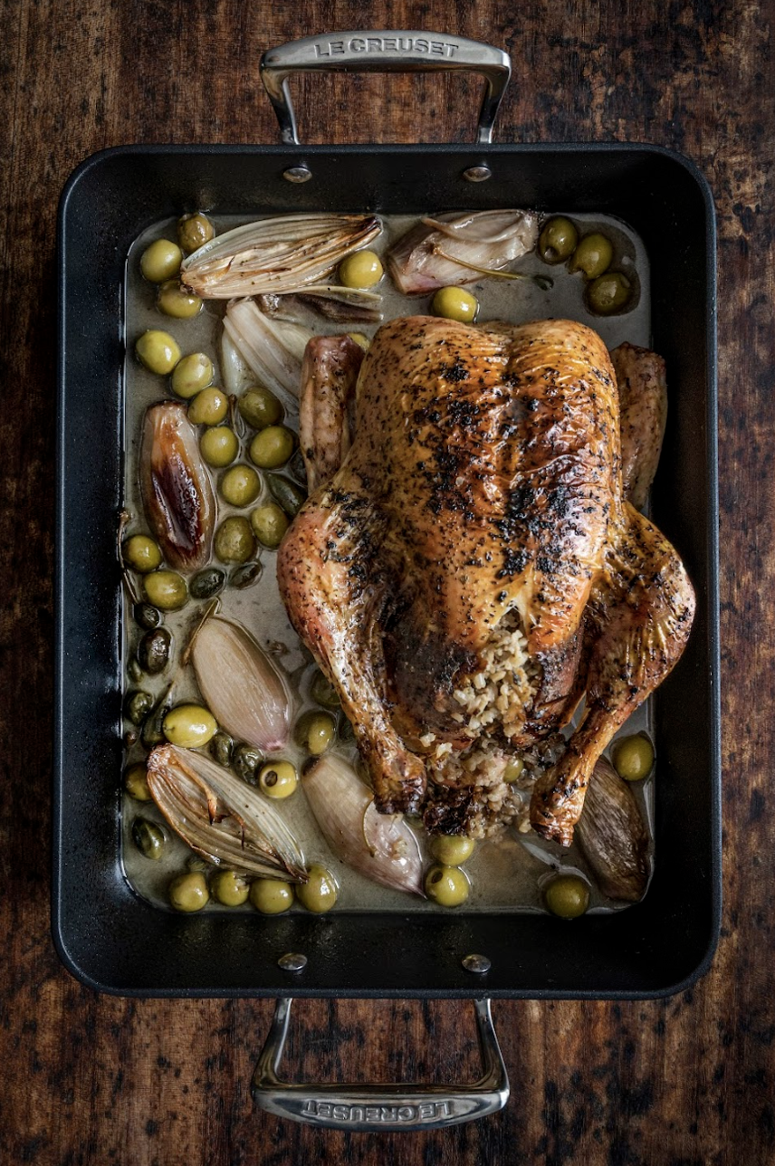 Whole roasted chicken in a roasting pan with garlic cloves and green olives, on a wooden surface.