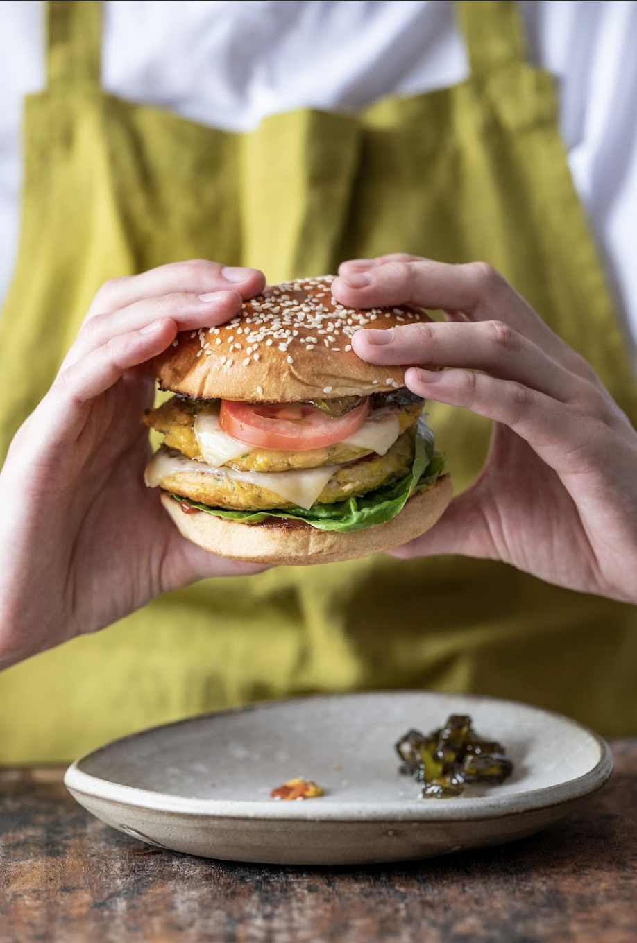 Person holding a sesame seed hamburger with lettuce, tomato, cheese, and a chicken patty, with pickles on a white plate in the background.