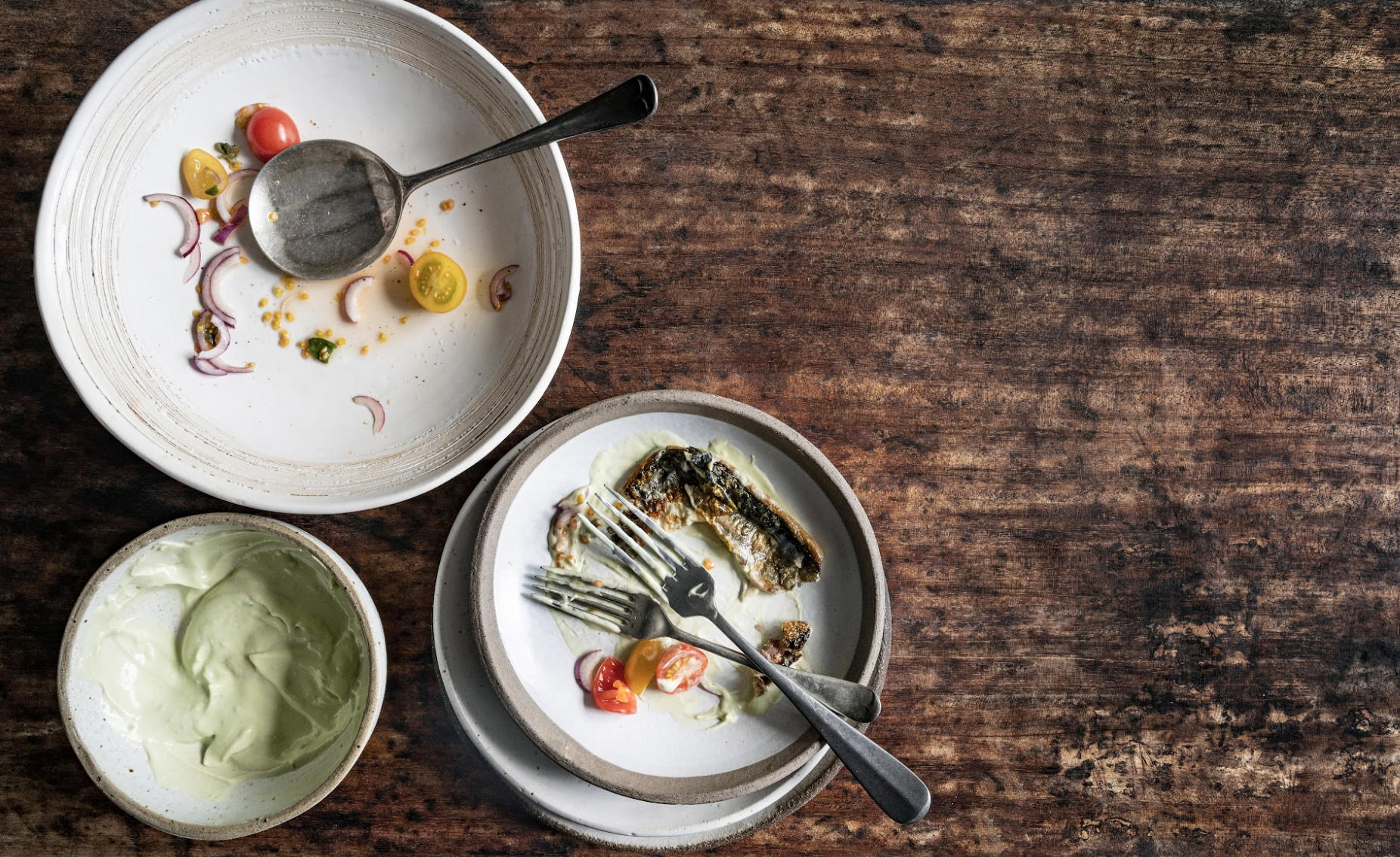 Empty plates with fish bones, sauce, and salad remnants, along with a bowl of avocado or guacamole on a wooden table.