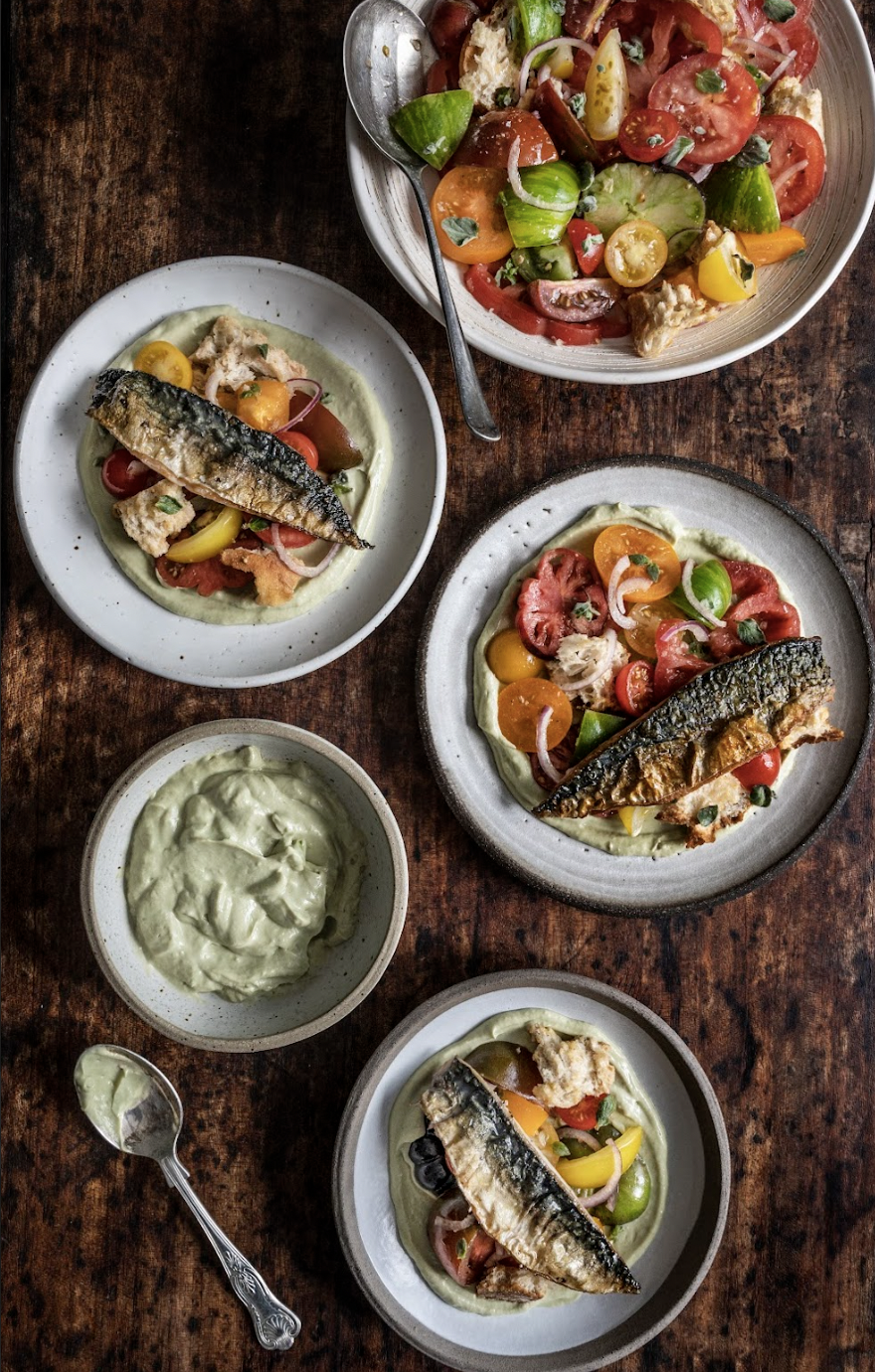 A rustic wooden table with three plates of grilled sardines on vegetable and tomato salads, a bowl of guacamole, and a large bowl of mixed salad with cherry tomatoes and greens.