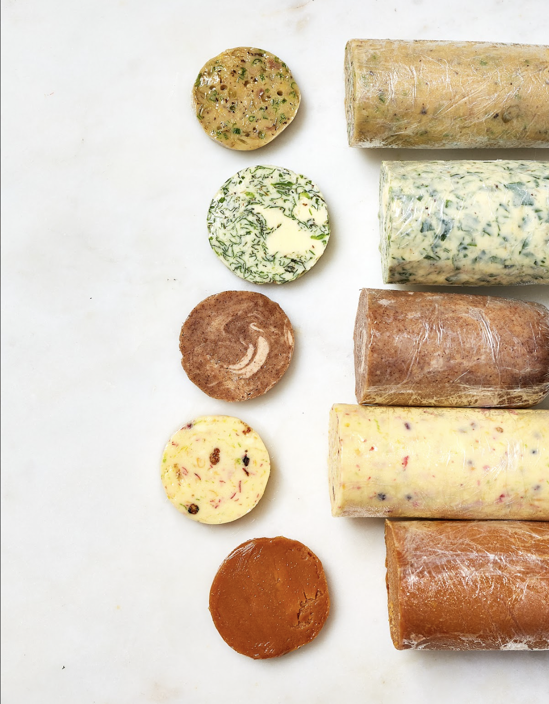 Various types of soap bars and rounds arranged vertically on a white surface, with some wrapped in plastic and others unwrapped, showcasing different colors and textures.