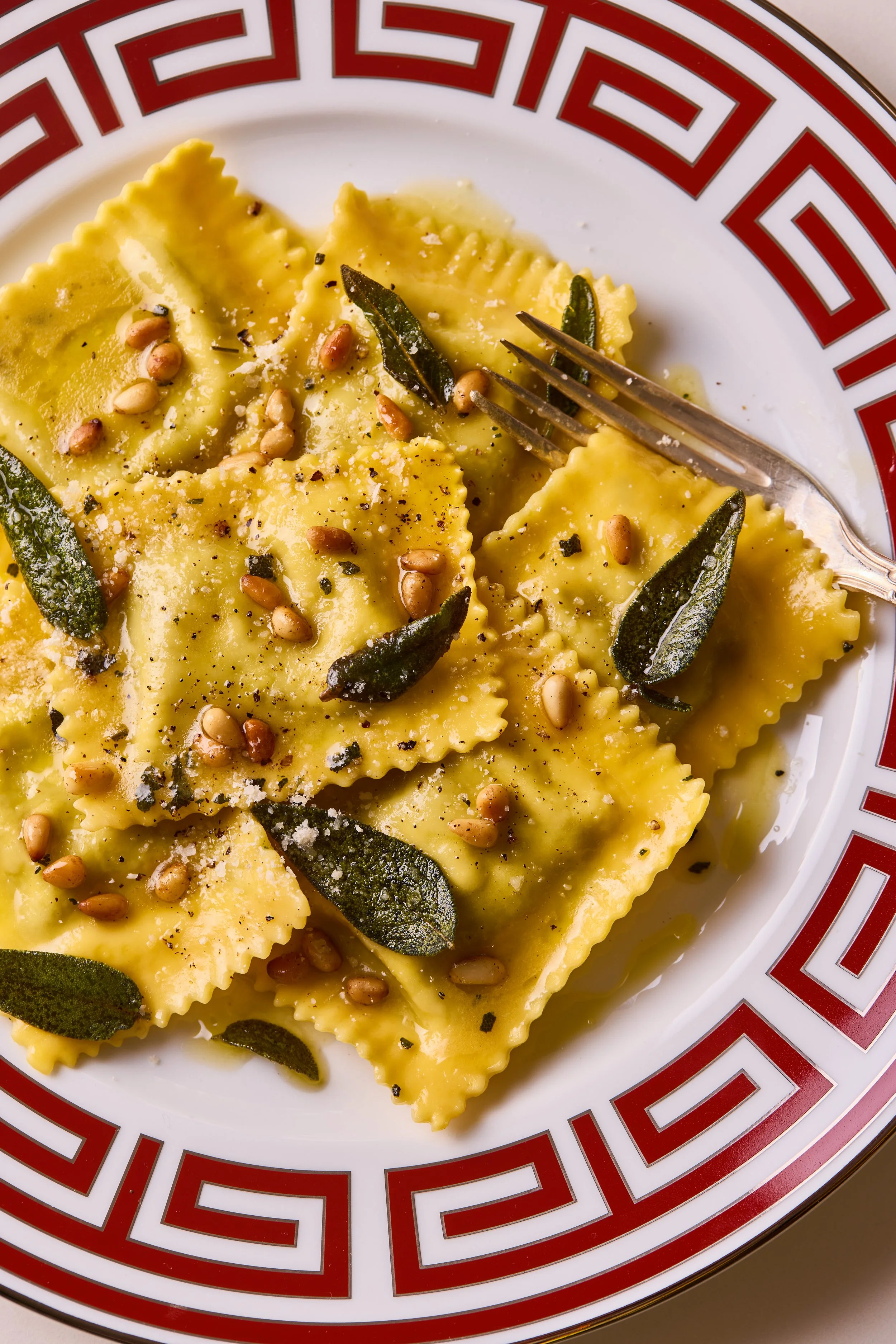 Pumpkin ravioli with crispy sage and hazelnuts from a top down angle. On a plate with red pattern trim.