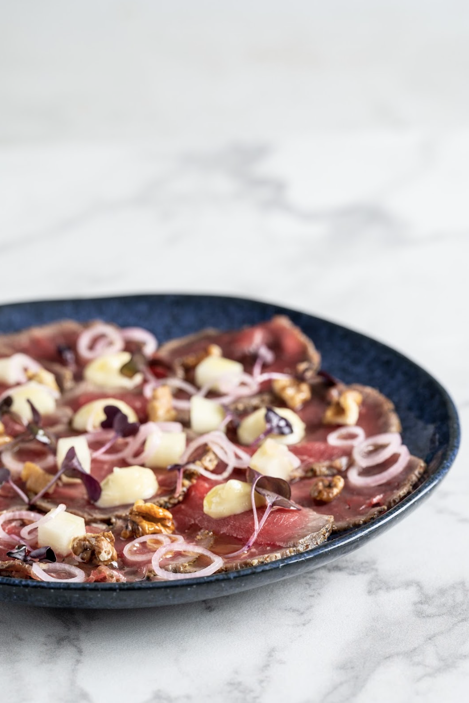 Thinly sliced cured meat garnished with chopped onions, microgreens, cheese cubes, and nuts on a dark plate.