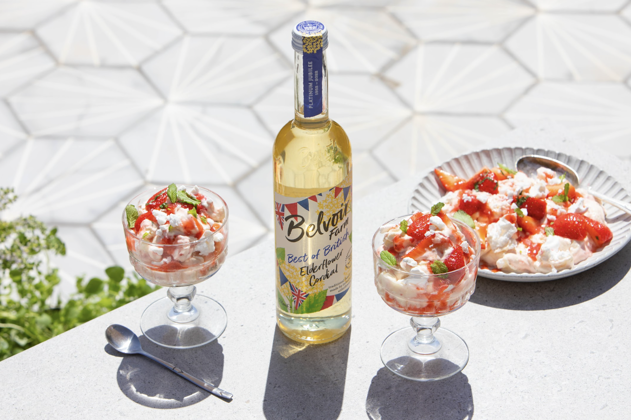 Two glasses of strawberry and cream dessert with mint garnish, a plate of the same dessert with strawberries and meringue, and a bottle of elderflower cordial on a white outdoor table with geometric patterned tiles in the background.