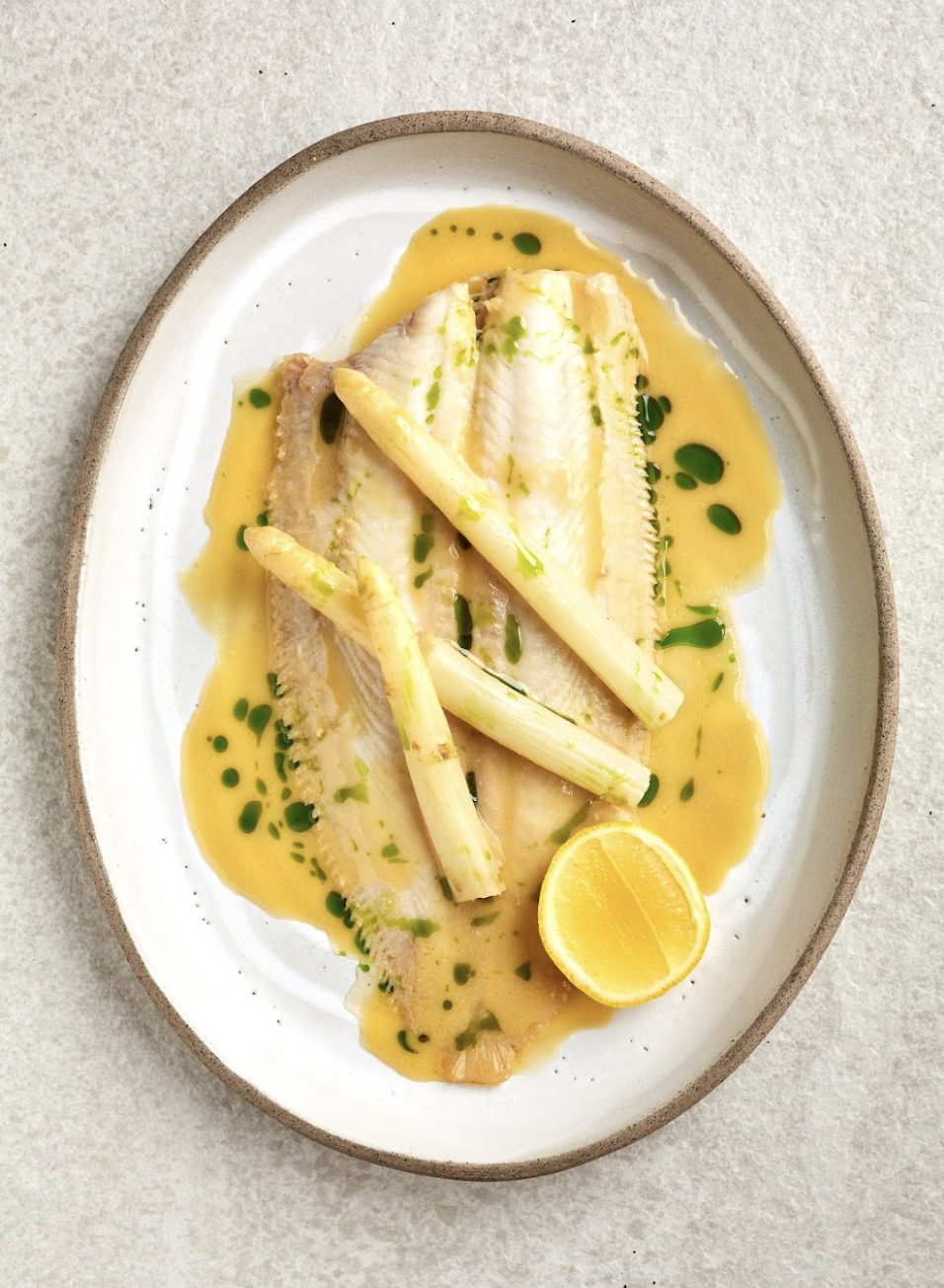 A cooked white fish fillet served with white asparagus, lemon wedge, and a yellow sauce with green droplets on a beige and white ceramic plate.