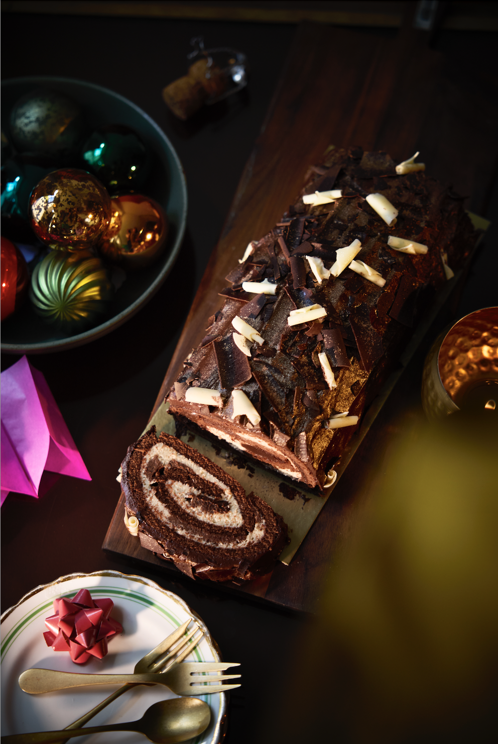 Waitrose chocolate swiss roll, top down shot. A colourful bowl of baubels and a small plate with a gold fork sit around it.