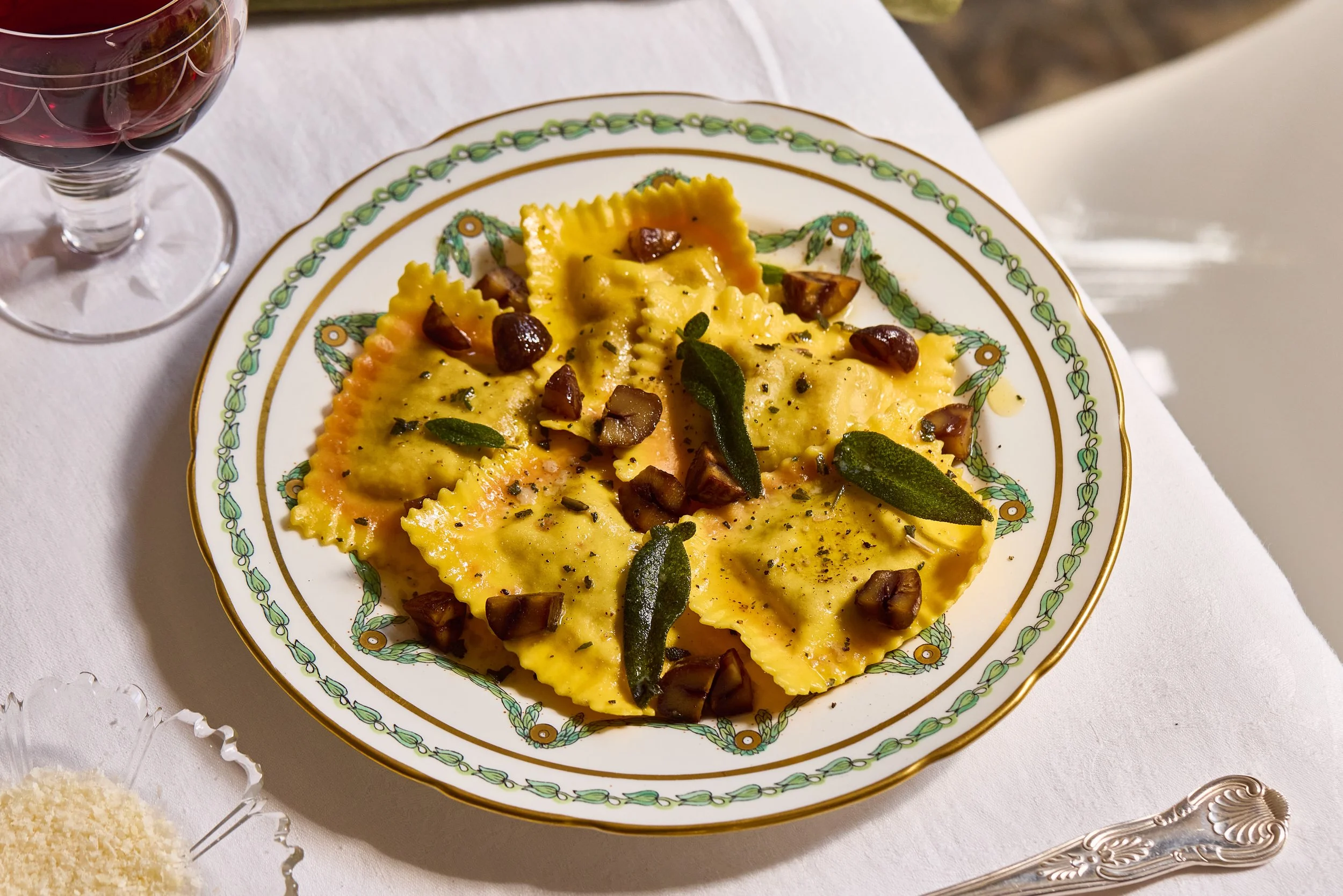 Pumpkin raviloli plated on a green trim plate. SHot from a low angle in dappled sunlight with Parmesan, a glass of red wine and cutlery around the edges of the images.