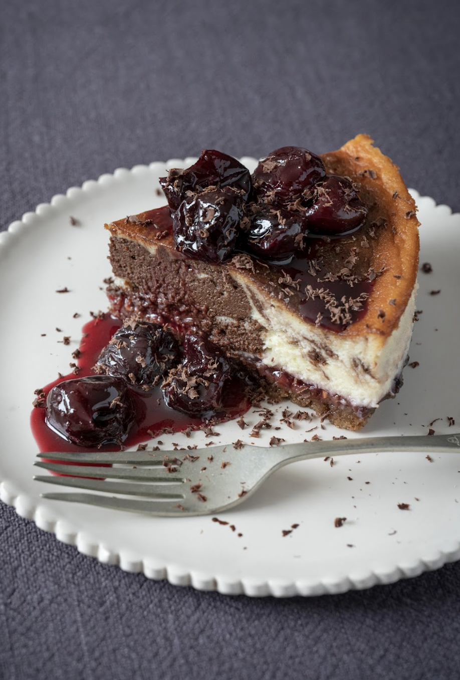 Slice of chocolate cherry cheesecake with cherry topping, on a white plate with a fork, against a dark background.