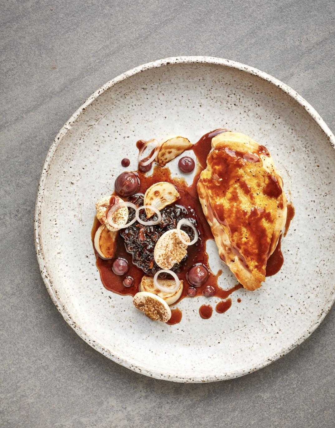 A plated gourmet dish with a stuffed pastry topped with sauce, served with fruit and garnished with sliced garlic and onions.