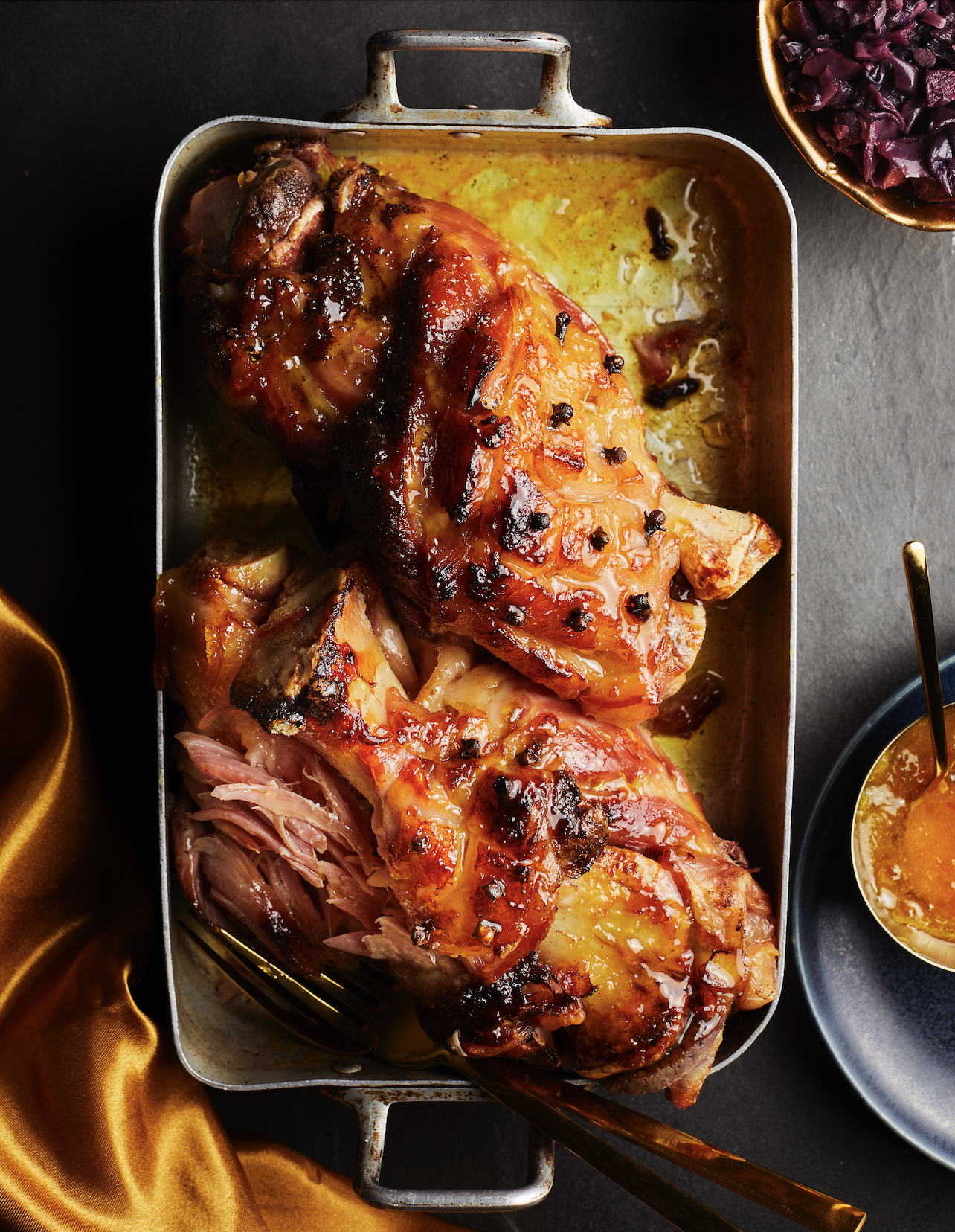 Baked ham with a caramelized glaze in a roasting pan, garnished with black peppercorns, alongside side dishes including diced purple cabbage in a bowl and some gravy in a small dish, on a dark surface with a gold cloth.
