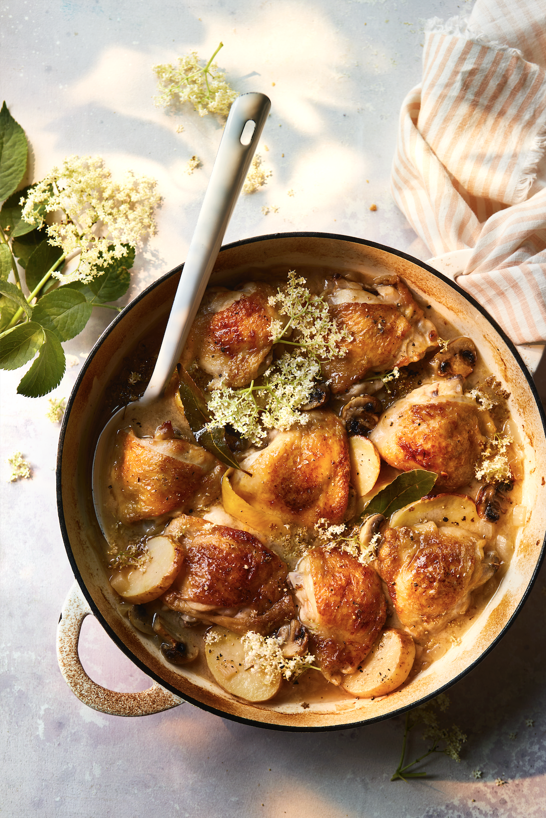A casserole dish with cooked chicken thighs and potatoes in a creamy sauce, garnished with herbs and flowers, on a light surface with a striped cloth nearby.