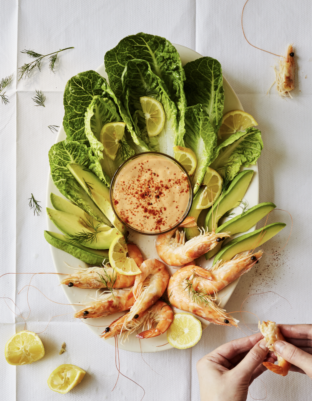 Seafood platter with shrimp, lemon slices, avocado, lettuce, and a dipping sauce in the center on a white tablecloth.