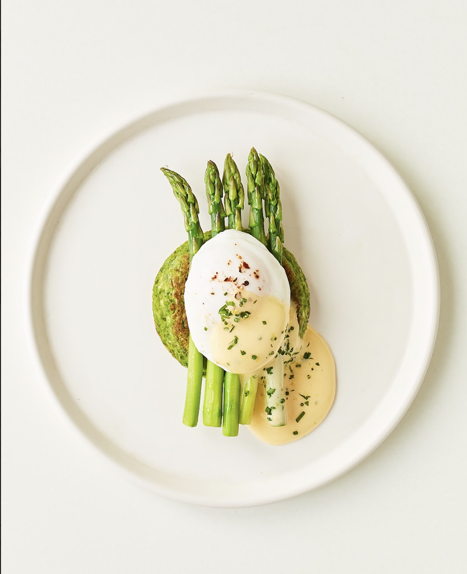Poached egg on asparagus with green vegetable patty and creamy sauce, garnished with herbs on a white plate.