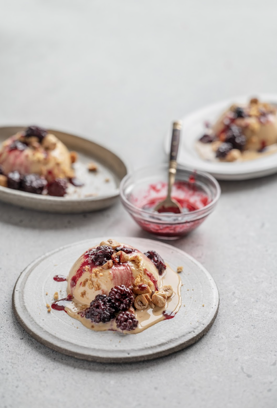 A plate of vanilla ice cream topped with blackberry sauce, blackberries, chopped nuts, and caramel sauce, with additional plates of ice cream and a small bowl of berry sauce in the background.
