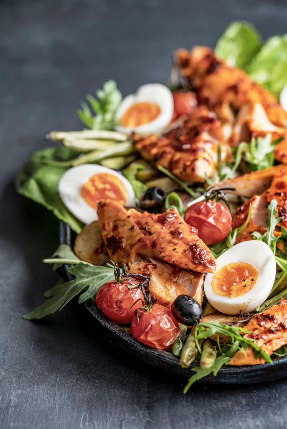 A fresh salad with grilled chicken, cherry tomatoes, halved soft-boiled eggs, mixed greens, and olives on a black plate.