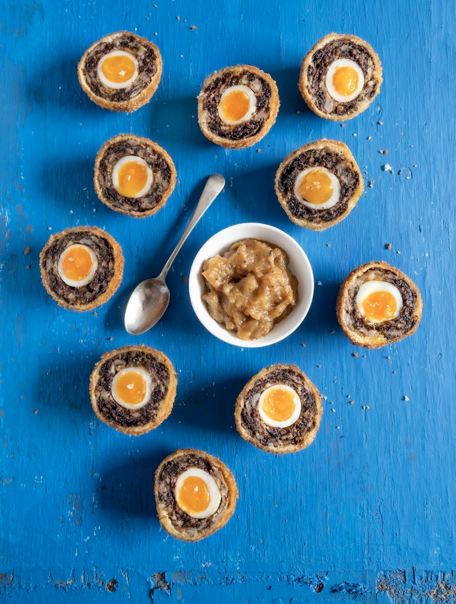 Nine Scotch eggs with soft boiled eggs in the center, arranged on a bright blue wooden surface around a small white bowl of musty or apple sauce and a spoon.