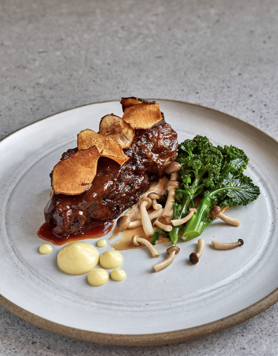 A plated dish featuring a piece of meat covered in barbecue sauce topped with potato chips, served with broccoli and mixed mushrooms, garnished with drops of yellow sauce.