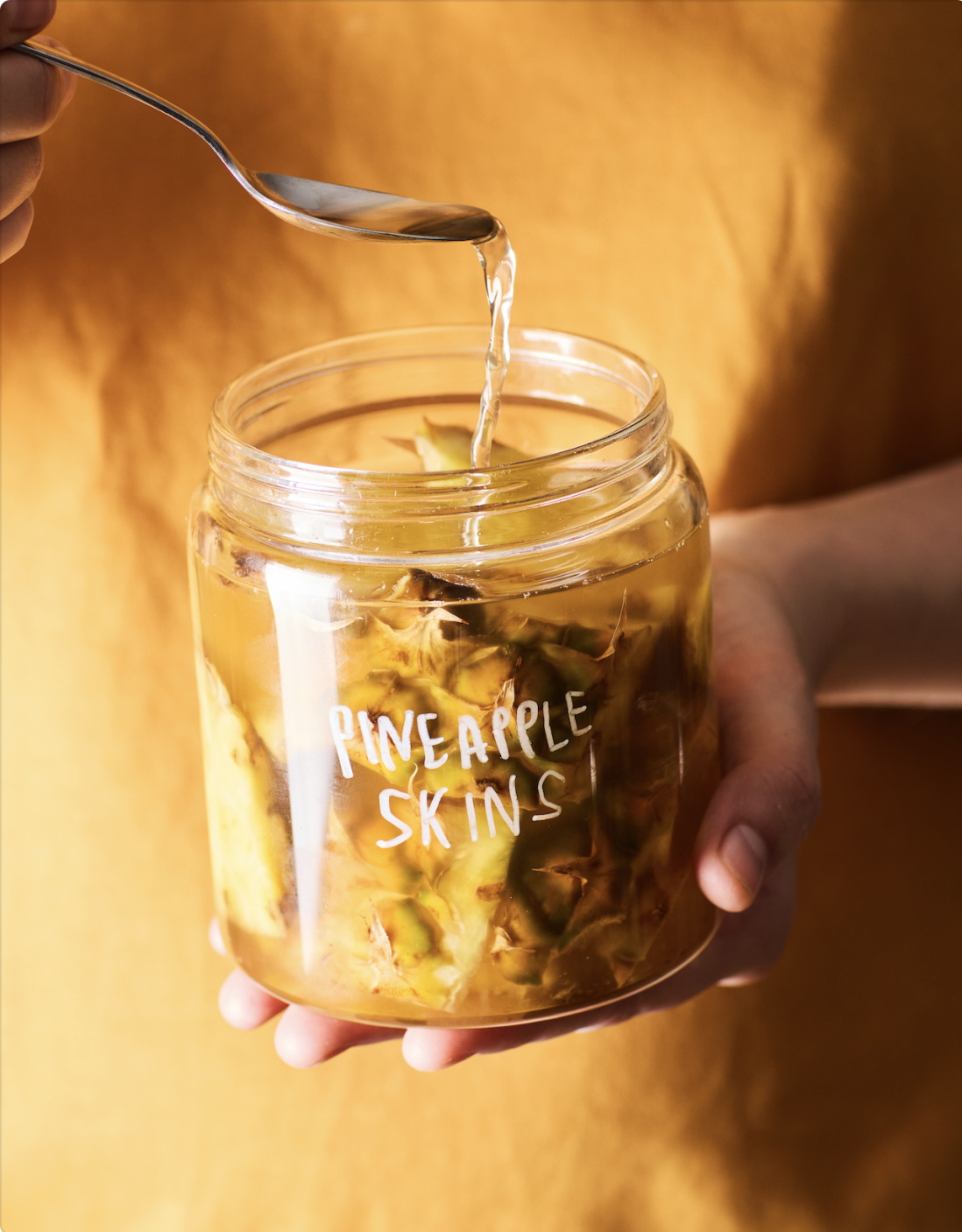 A person holding a glass jar with pineapple chunks and liquid, with the words 'Pineapple Skins' written on the jar. The person is pouring liquid into the jar from a spoon.