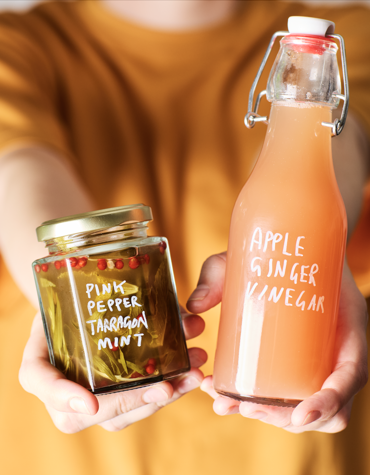 A person holding two glass bottles of infused drinks; one labeled 'Apple Ginger Vinegar' and the other 'Pink Pepper Tarragon Mint' with ingredients visible inside.