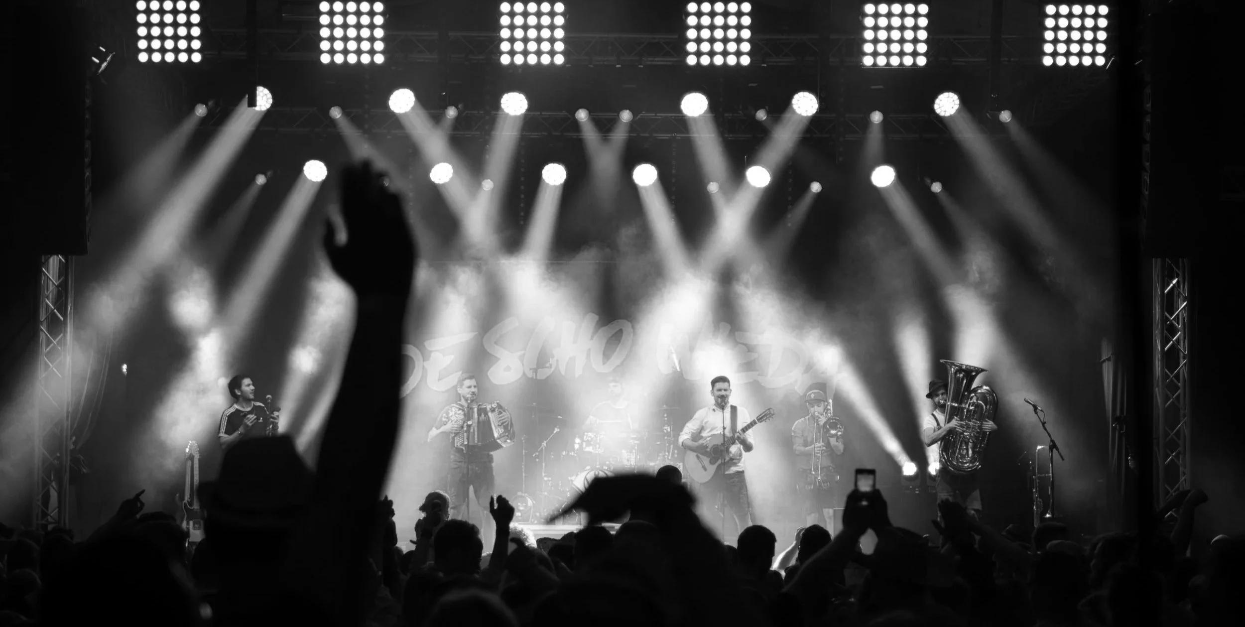 Schwarz-Weiß-Foto einer Bühne mit Musikern, hell beleuchtet, Publikum im Vordergrund, eins hebt die Hand mit Daumen nach oben.