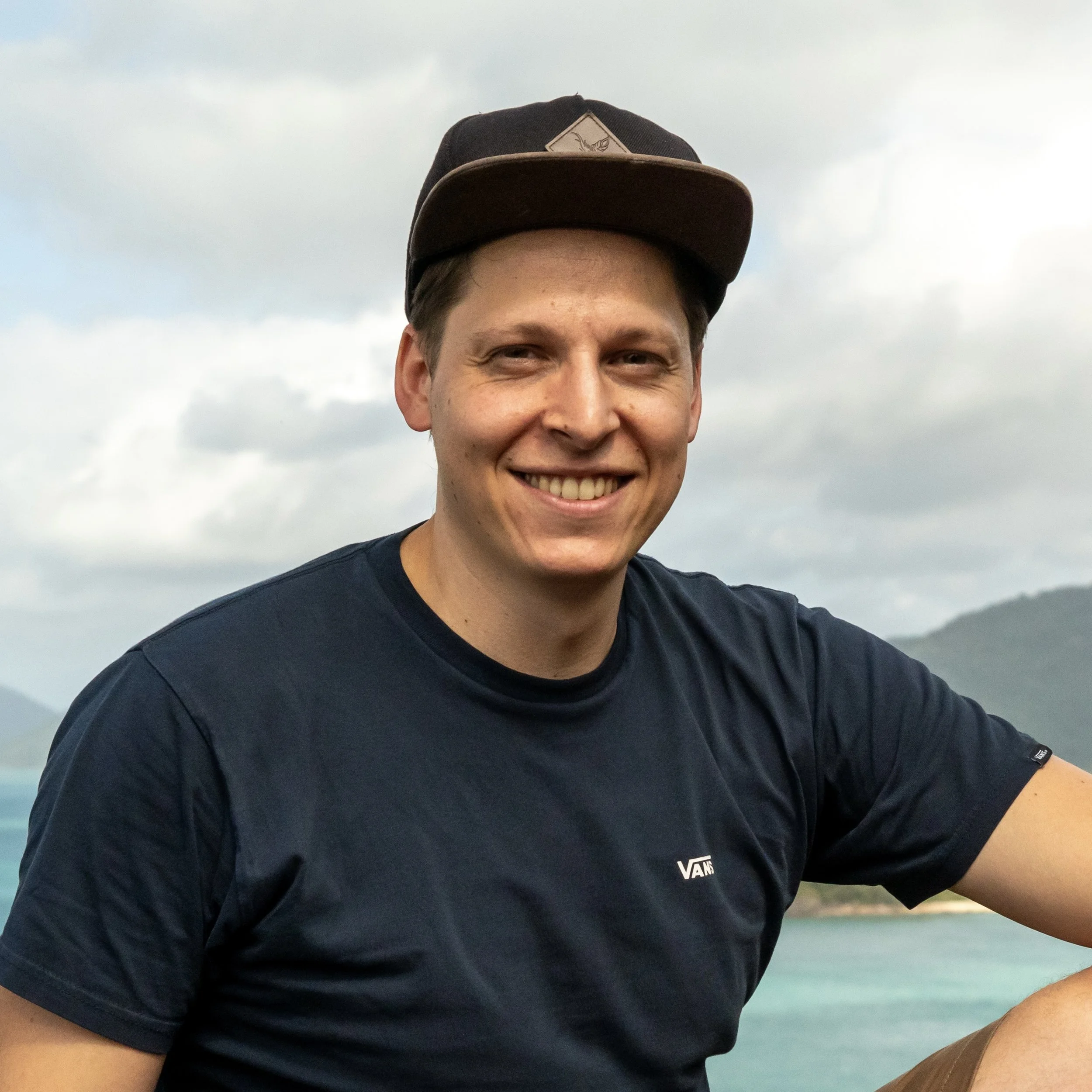 Ein lächelnder junger Mann trägt einen schwarzen Baseball-Cap und ein schwarzes T-Shirt vor einer naturnahen Kulisse mit Wasser und Hügeln.