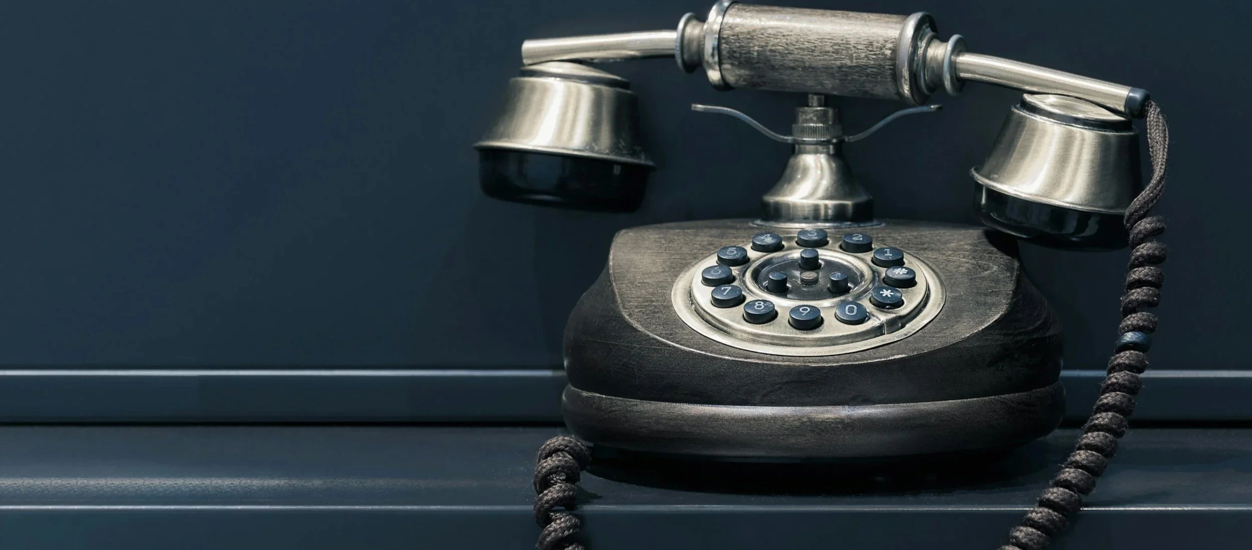 Alter curvasguter Telefon aus schwarzem Holz mit silbernen Metallteilen, Drehknöpfen und Kabeln, auf dunklem Hintergrund.