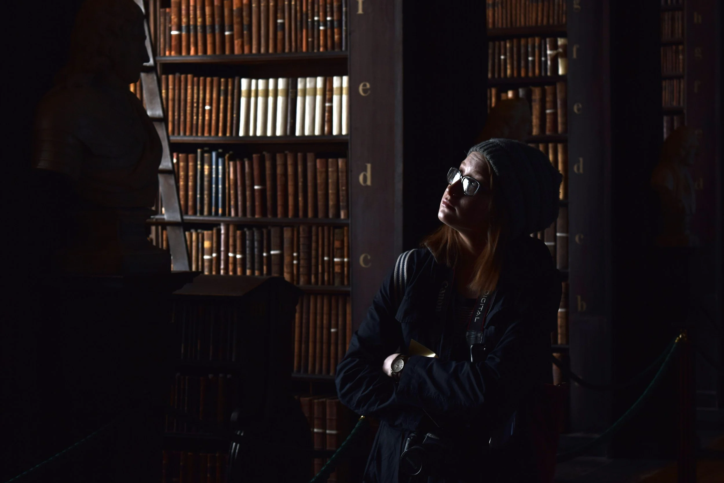 A woman with glasses and a beanie looking up in a dimly lit library with tall bookshelves filled with old books.