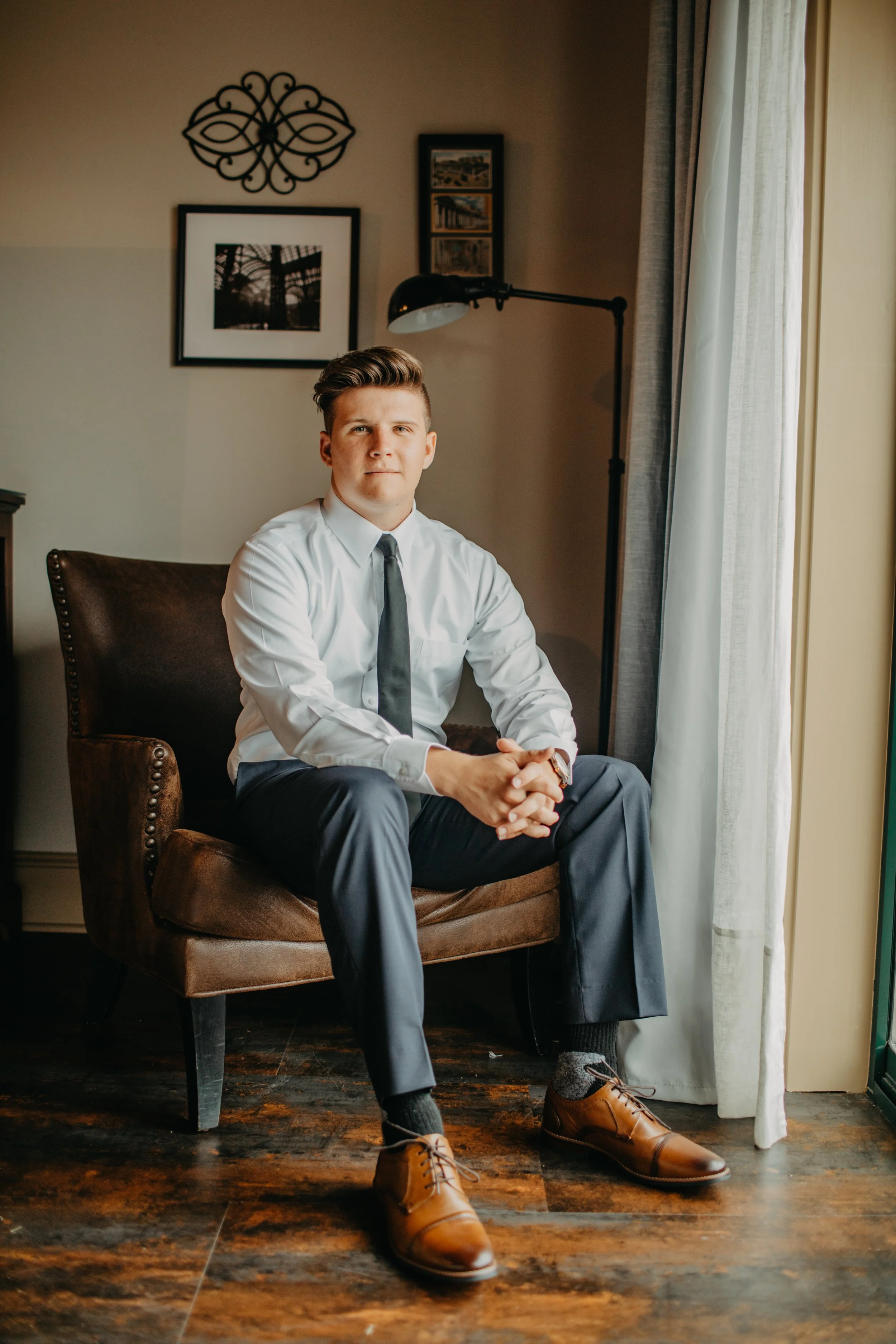 A young man in a white shirt, black tie, and gray trousers sitting on a brown armchair in a room with framed pictures on the wall, a black floor lamp, and a window with light-colored curtains.