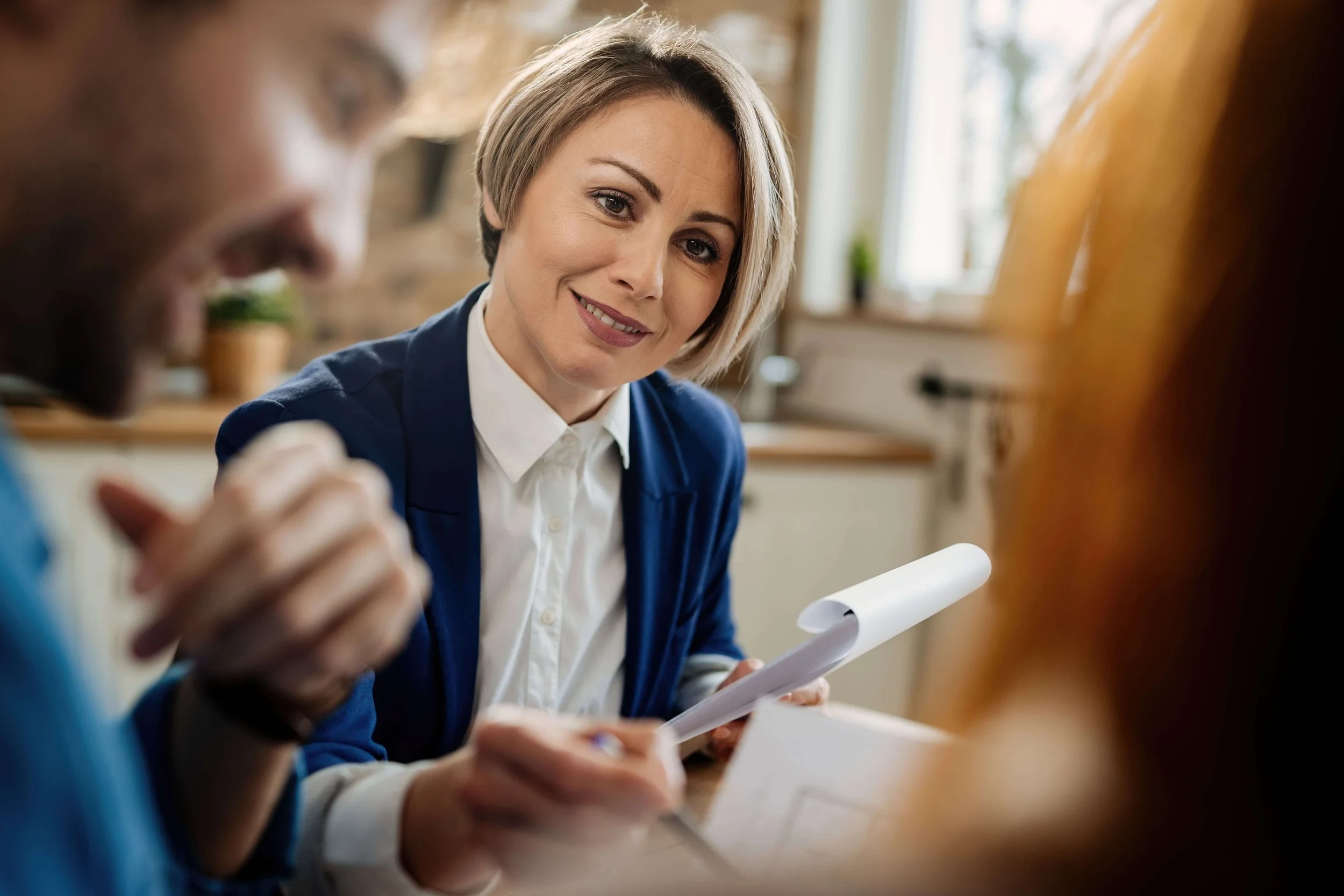 A woman with short blonde hair, wearing a blue blazer and white shirt, smiling while holding a notepad during a discussion with a man and a woman in a cozy, well-lit kitchen.
