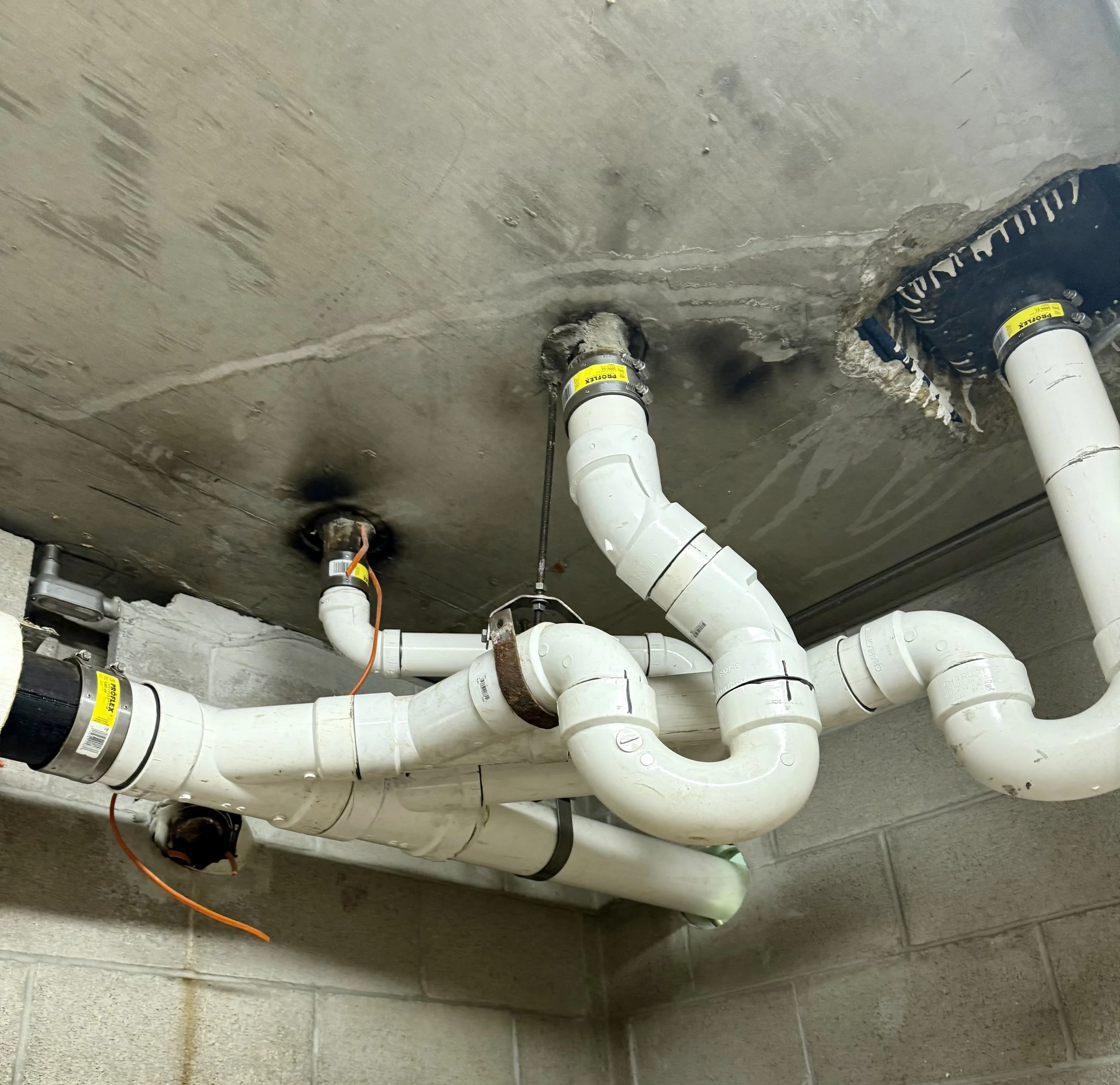 Underground plumbing pipes with various elbows and connectors, secured to a concrete ceiling with some insulation around the pipe openings.