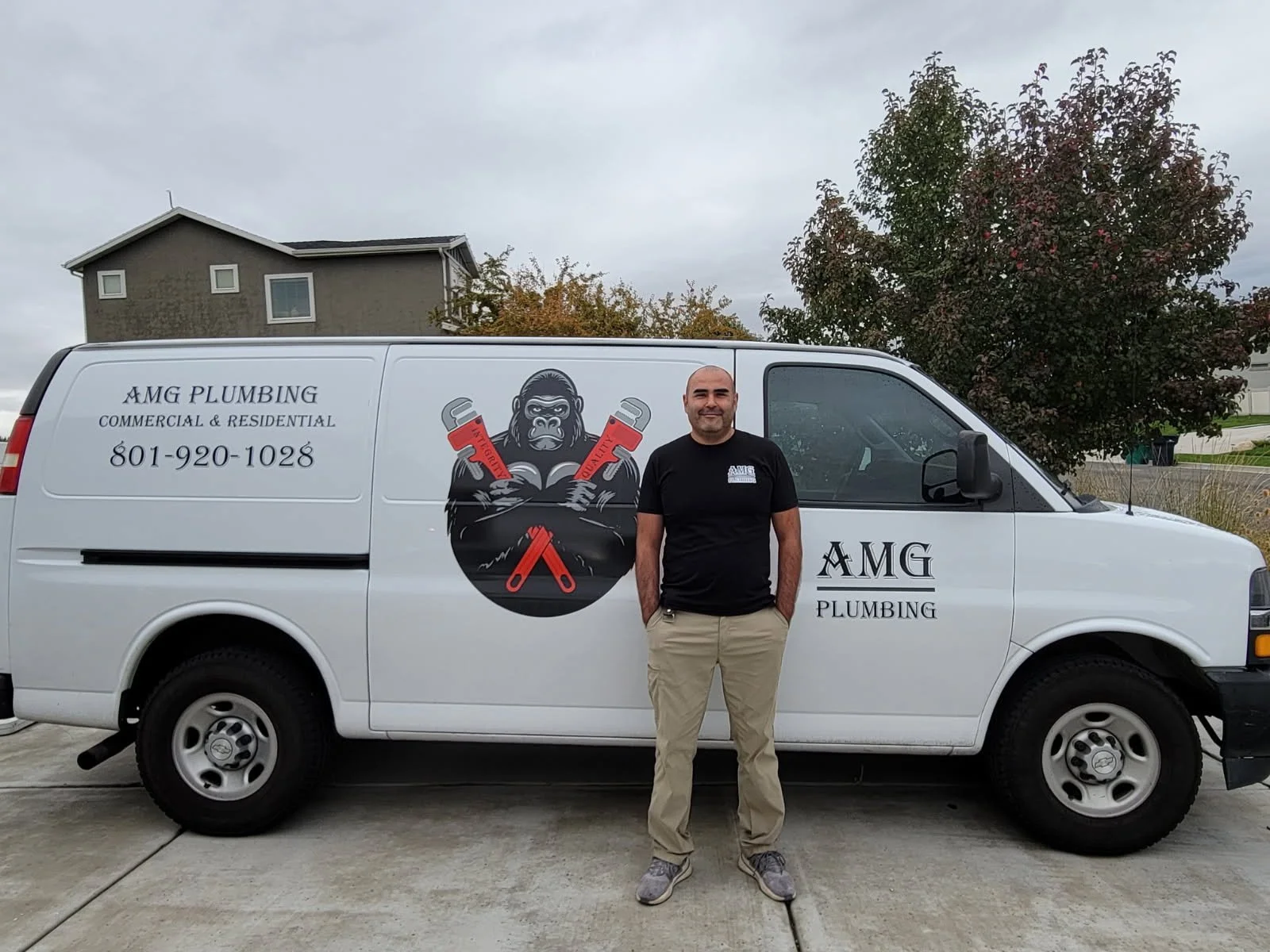 Man standing in front of a white plumbing service van with logo of an ape holding red pipes, and text "AMG Plumbing," with a background of residential homes and trees.