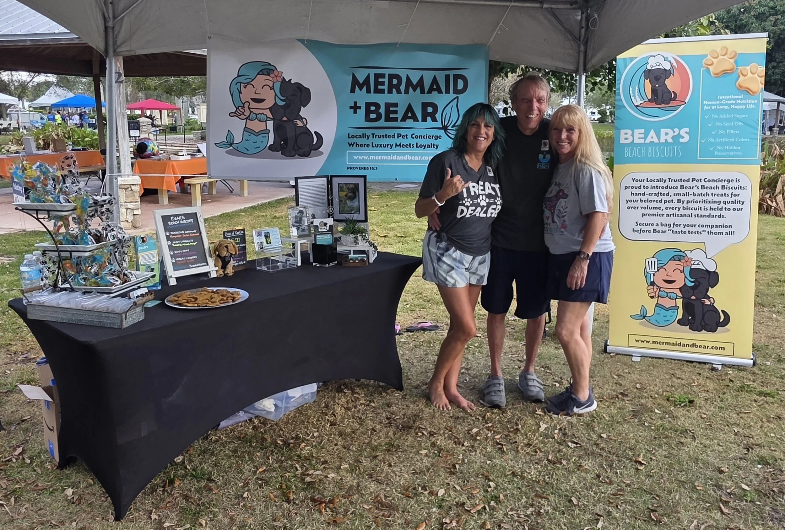 One man stands between two ladies smiling in front of signs and a table advertising Mermaid and Bear.