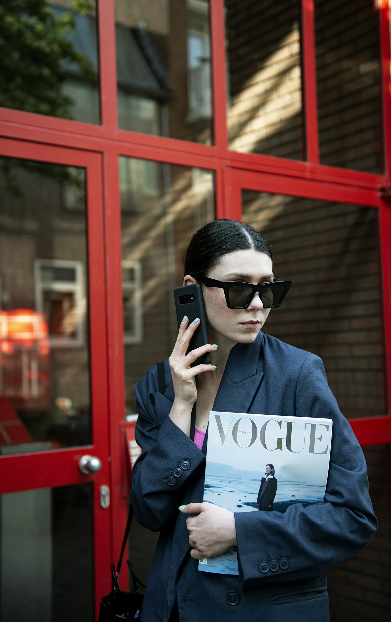 model photoshoot, Izabela Bartnicka Photography, model posing with Vouge magazine, wearing sunglasses, in front of glass door with red frames, fashion photoshoot