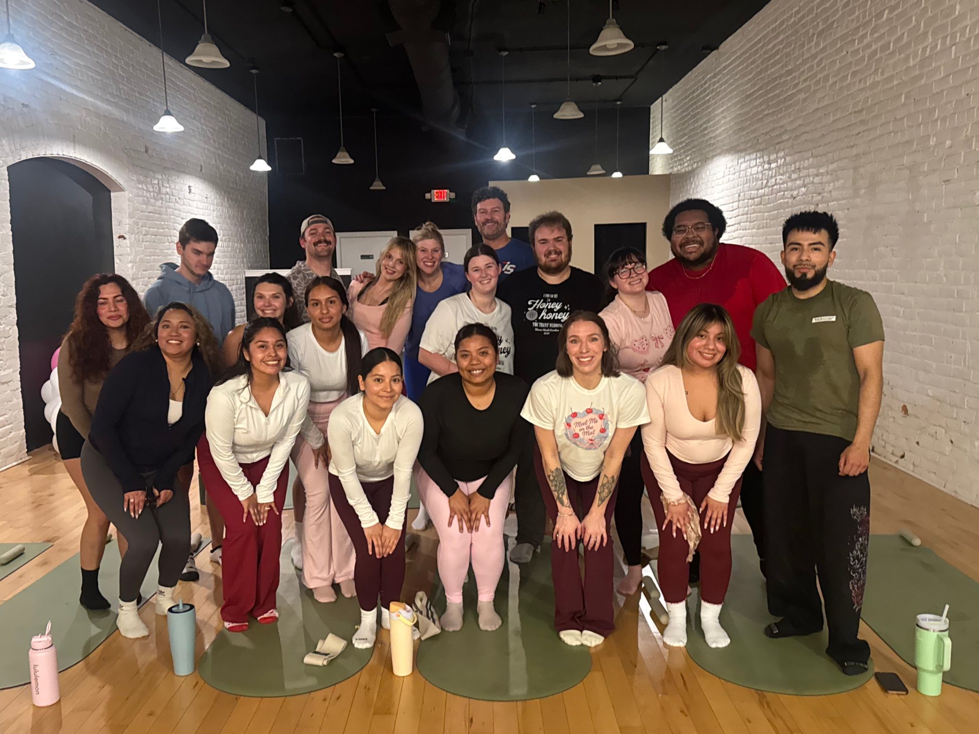 A group of people posing together in a room with white brick walls and wooden floors, some in casual wear and some in workout attire, suggesting a fitness or yoga class setting.