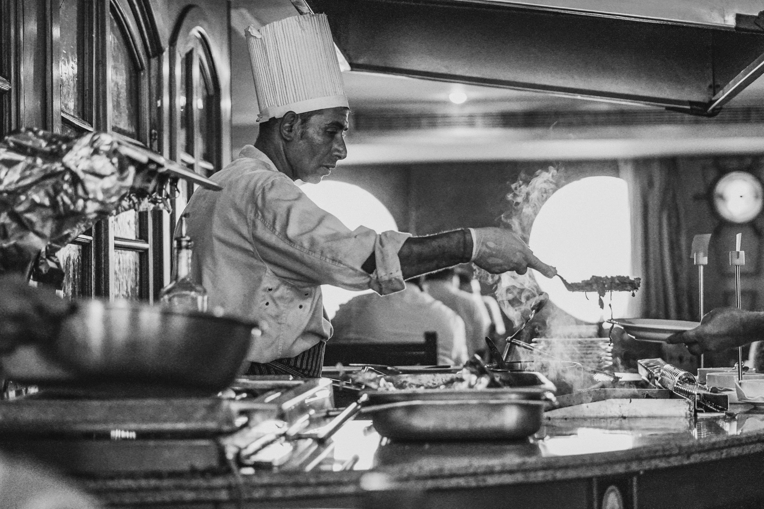 A chef in a tall hat and apron is cooking on a stove, using tongs to handle grilled food with steam rising, in a professional kitchen setting.