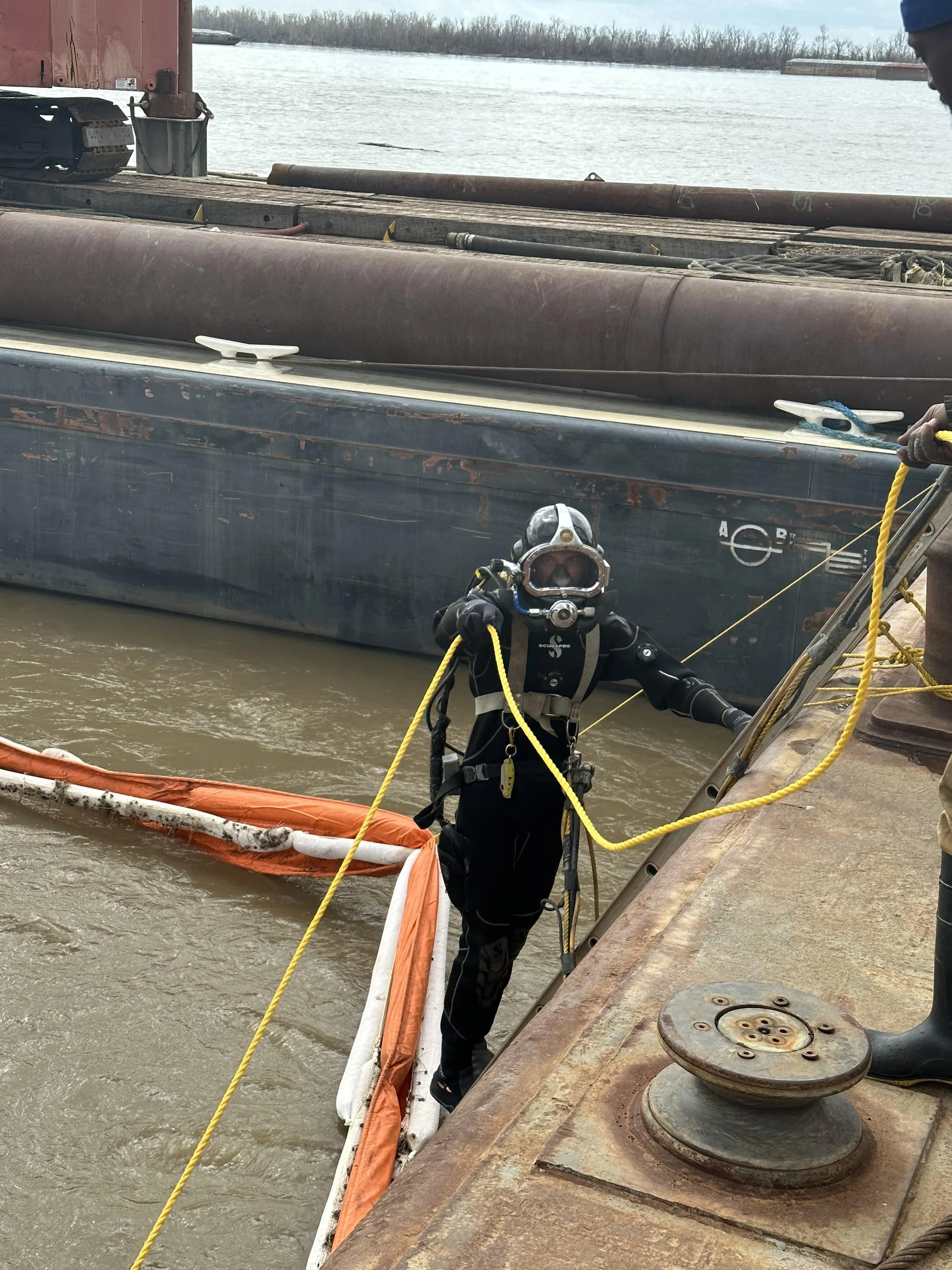 A diver in full gear standing on a rusty ship's deck, holding a yellow rope, near a submerged orange and white net in murky water, with large ships and water in the background.
