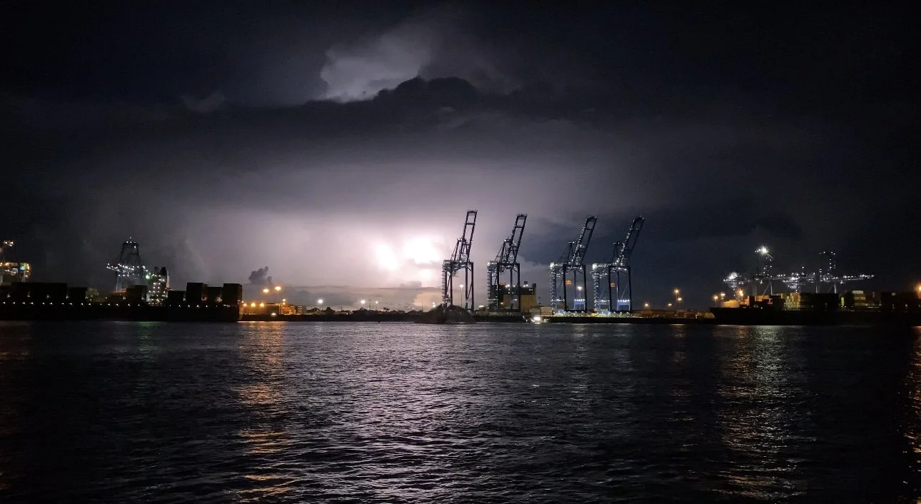 Nighttime view of a port with four large dock cranes illuminated, storm clouds overhead, and lightning in the distance, reflected on the water.