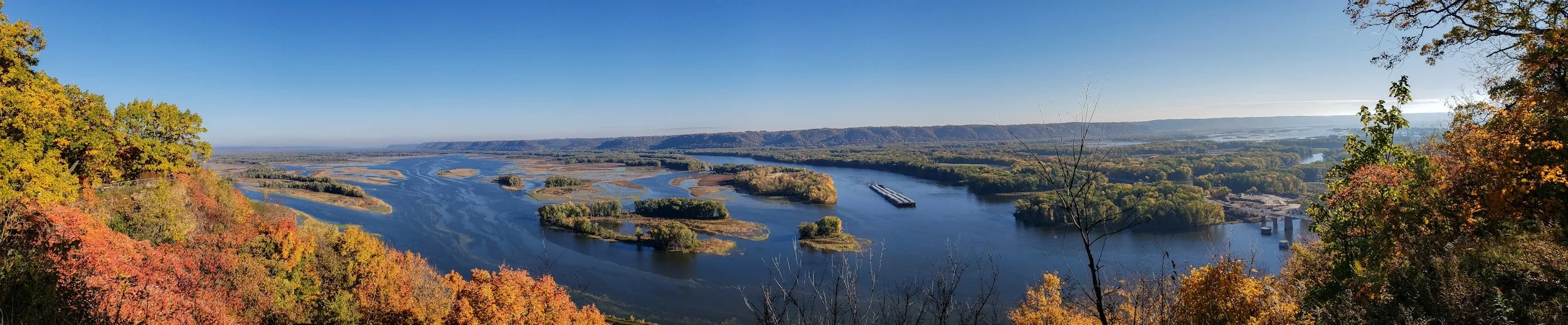 Panoramic view of a river winding through a landscape with colorful autumn trees in the foreground and distant hills under a clear blue sky.