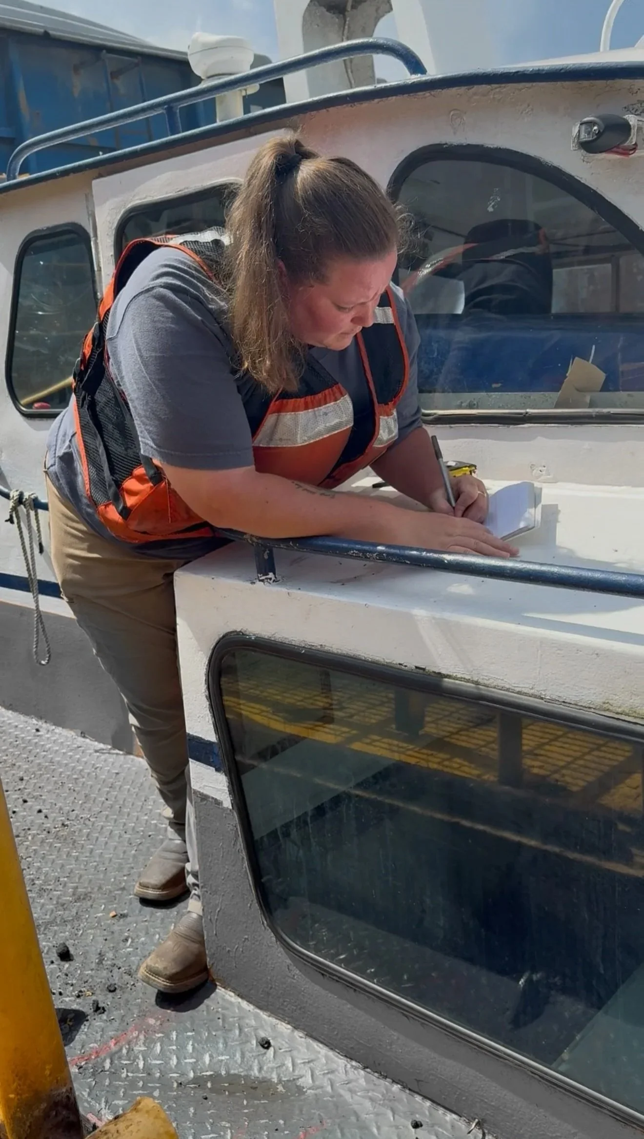 A woman wearing a reflective safety vest and tan pants is writing or drawing on a white surface of a boat, leaning forward with her left hand steadying her work.