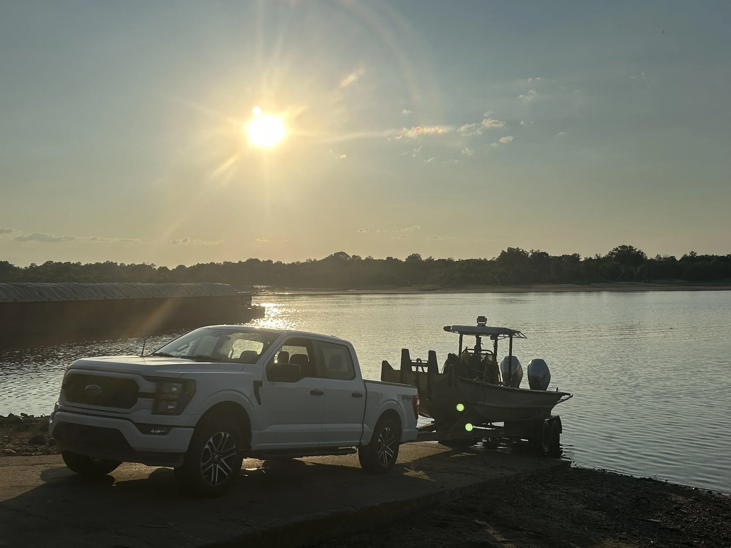 A white pickup truck parked near a body of water, towing a small motorboat on a trailer, with the sun shining in the sky and reflecting on the water surface.
