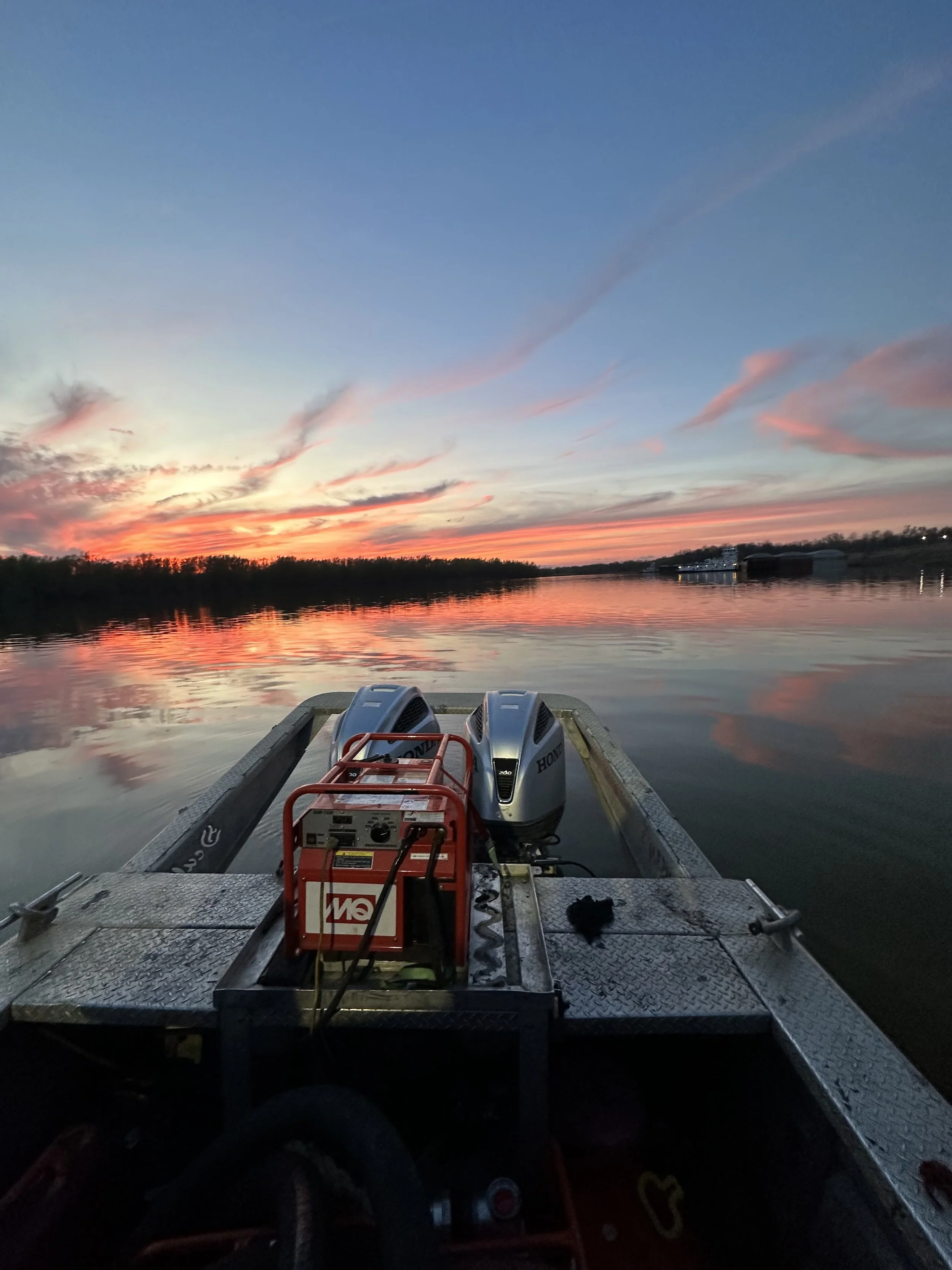 A boat on a river during sunset with colorful clouds in the sky and the water reflecting the sky and surrounding landscape.