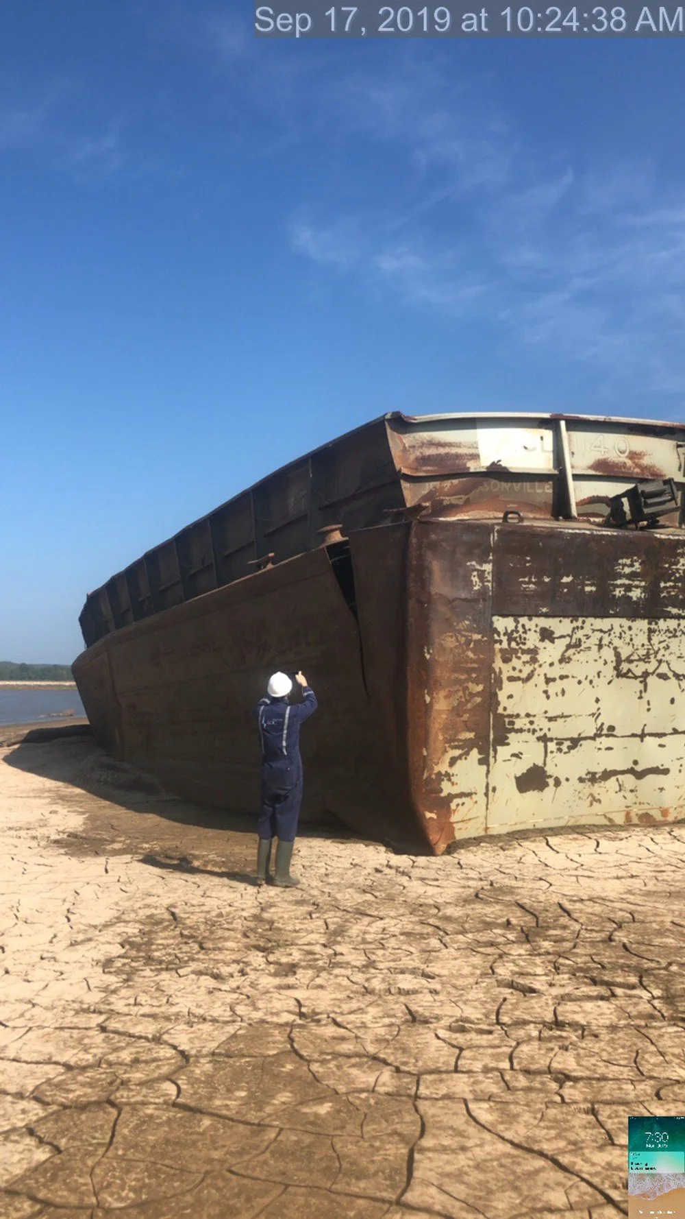 A person wearing a hard hat and boots taking a photo of a large, rusted abandoned boat on dry cracked land under a partly cloudy sky.