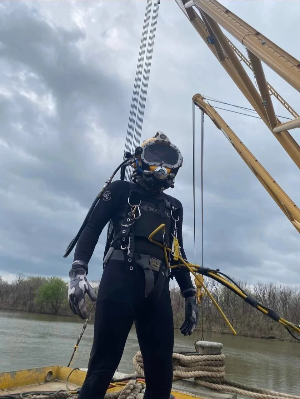 A person in a deep-sea diving suit with a helmet, standing on a boat near a body of water, with a crane in the background.