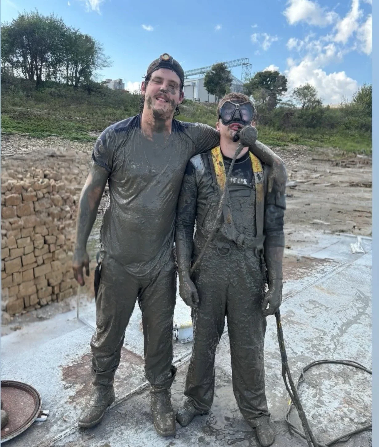 Two men covered in gray mud, standing outdoors, with one wearing a headlamp and wearing a helmet with a mask, and the other with a headlamp on his head. They are smiling, and the background shows a partly cloudy sky, green trees, and a concrete area.
