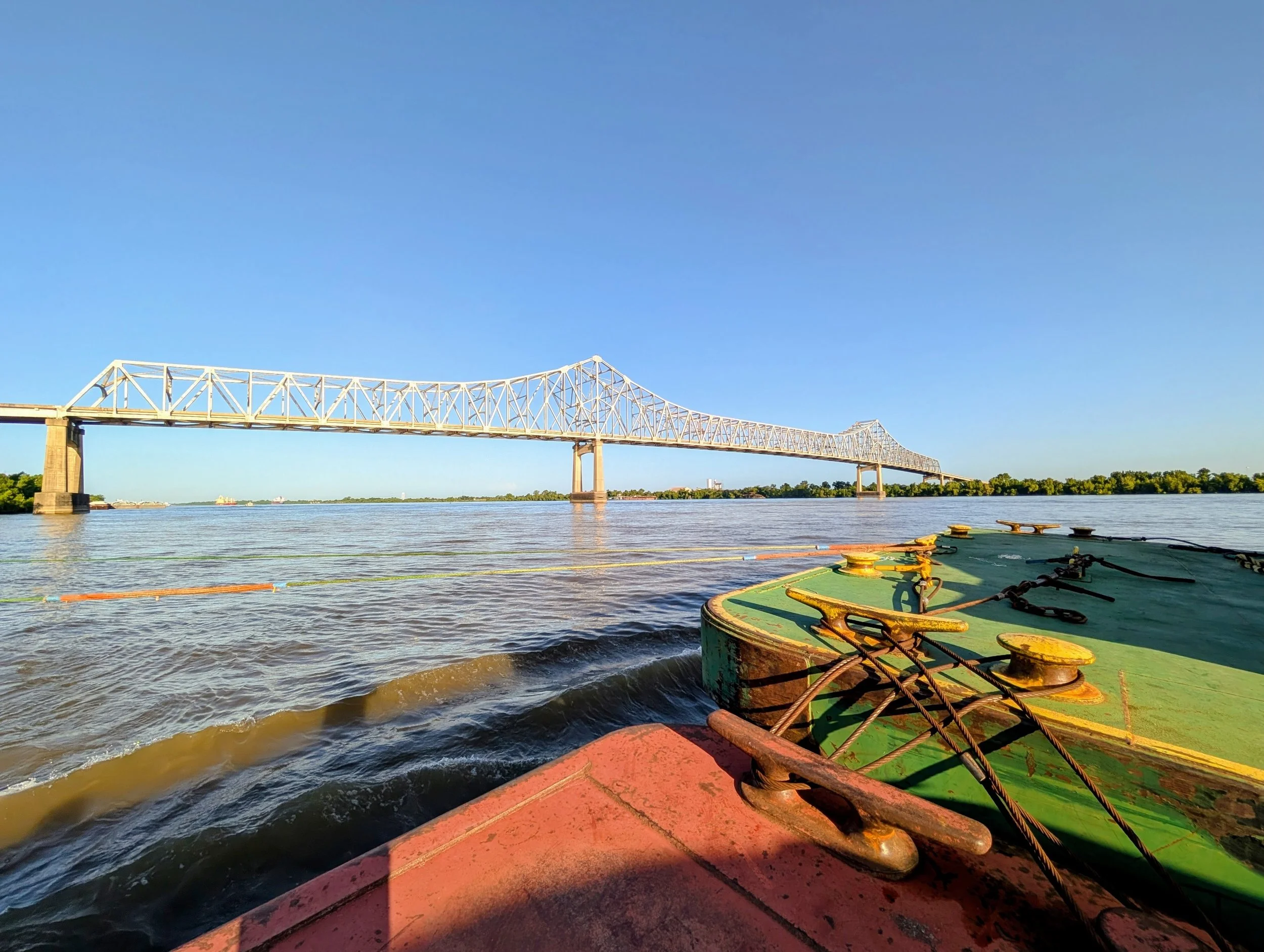 View of a bridge over a river with a boat in the foreground, with parts of the boat visible including ropes and pulleys, under a clear blue sky.