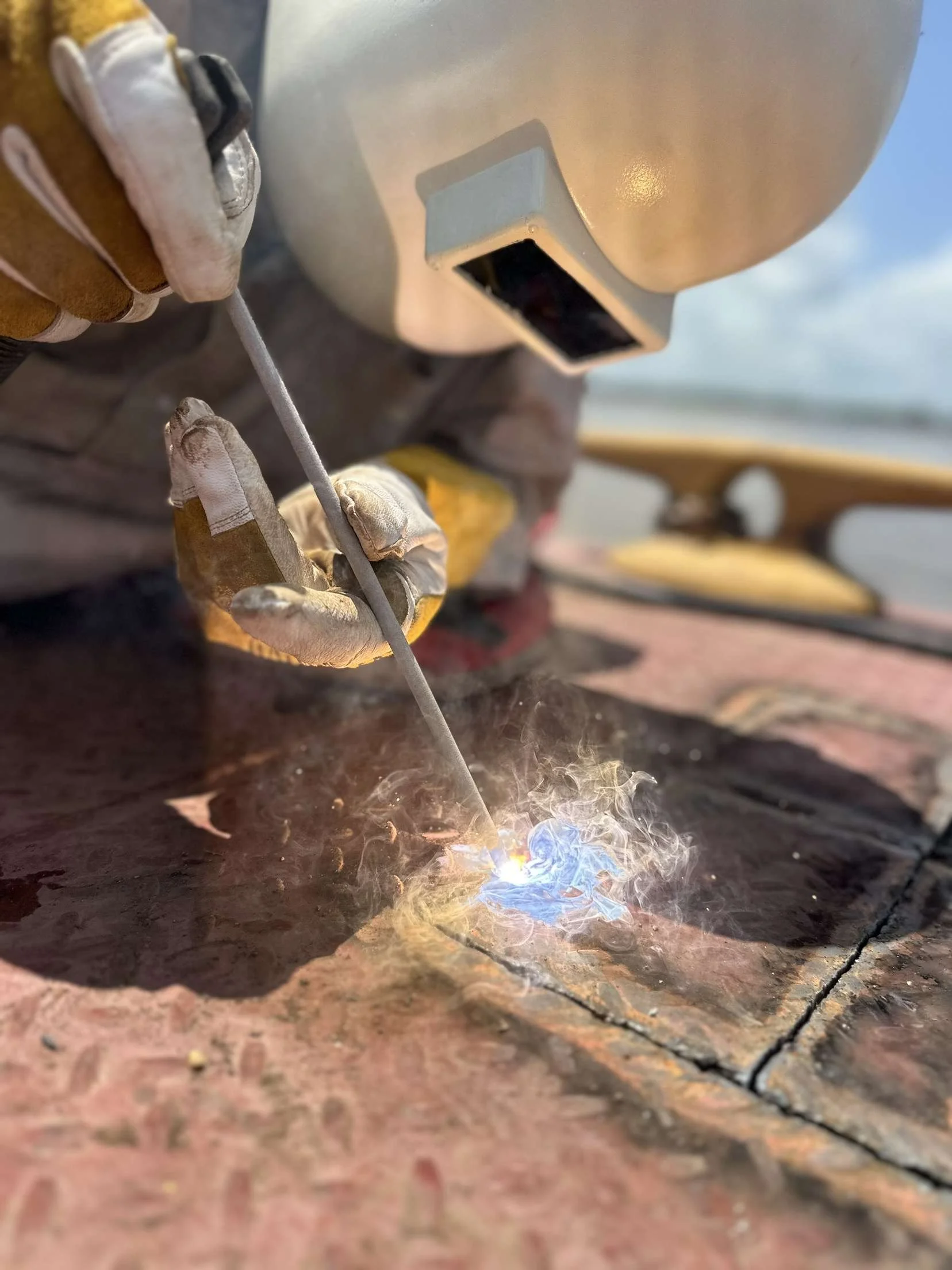 A person welding metal on a rusty surface, wearing protective gloves and a welding helmet, with sparks and smoke from the welding process visible.