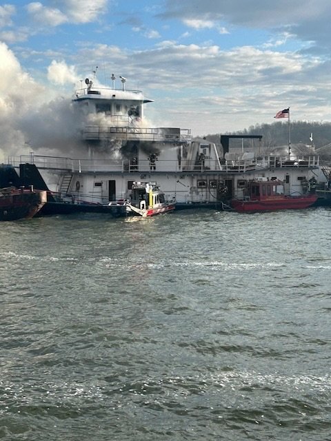 Fire on a large white boat with smoke billowing from the upper deck, two smaller boats nearby, and an American flag flying on a pole.