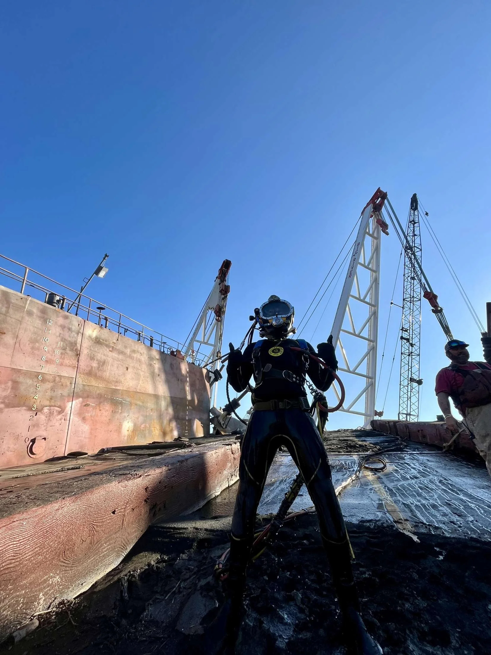 A person wearing safety equipment and an astronaut suit with a helmet is standing on a wet surface at a construction site, with large cranes and a partly cloudy blue sky in the background.